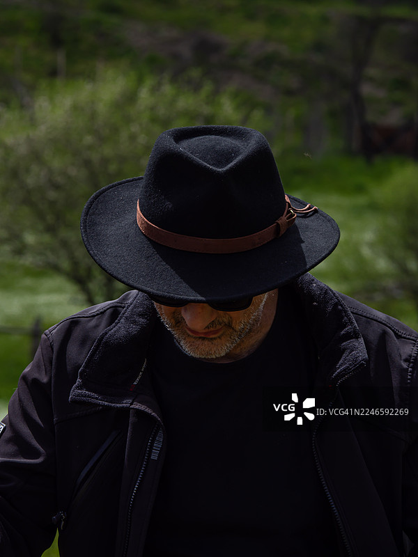留着胡须的男子戴着黑帽、穿着夹克，站在户外的绿色背景前图片素材