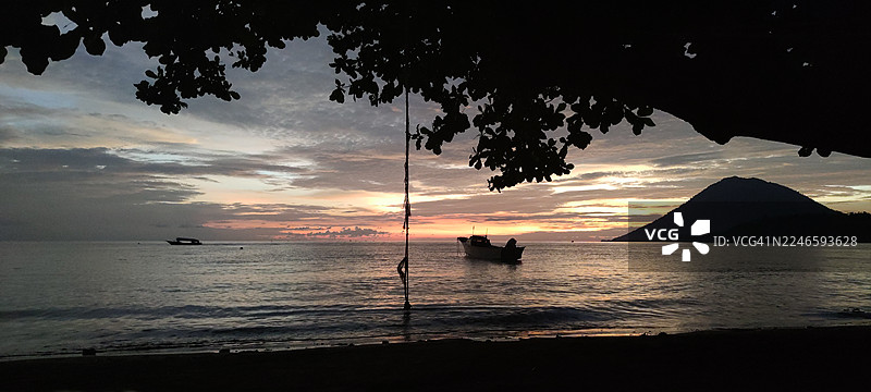 海边树的剪影，映衬着色彩鲜艳的日落天空，有船只和远山，布纳肯，印度尼西亚图片素材