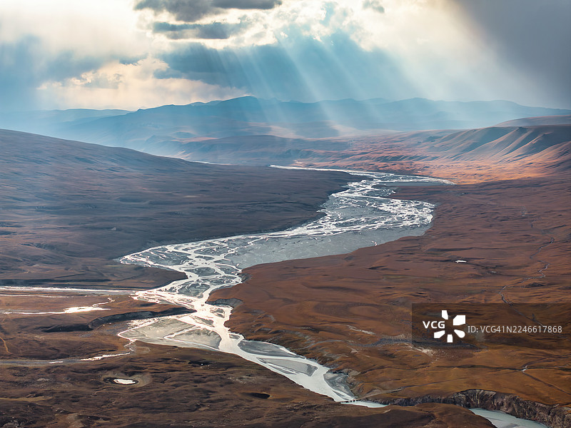 吉尔吉斯斯坦天山山脉,广阔干旱的高原上,一条宽阔的辫状冰川河切割着大地,在多云的天空下,阳光穿透云层,呈现出戏剧性的空中视角。图片素材