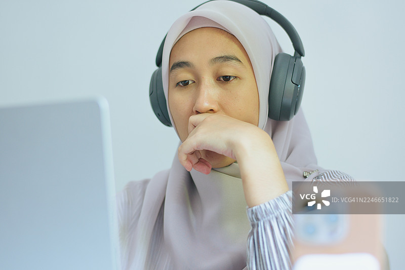 戴着头巾、使用笔记本电脑和耳机的专注的穆斯林女性在家办公图片素材