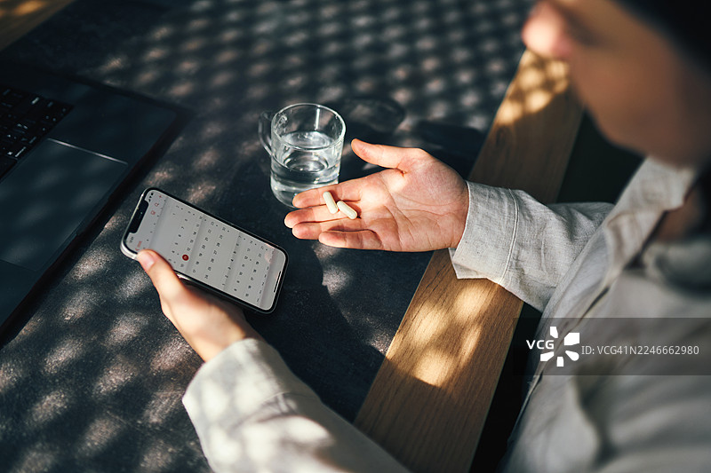 一个人在笔记本电脑和水杯旁，用智能手机查看日历，手里拿着药丸，办公室桌景图片素材