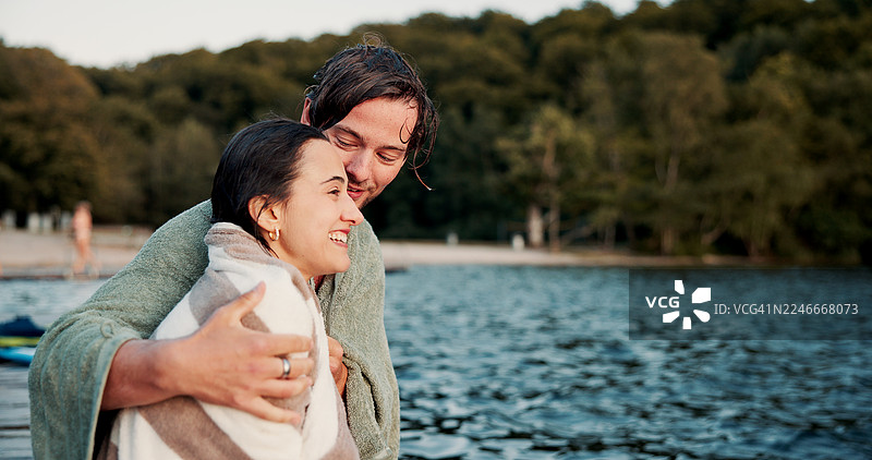 情侣在假期里依偎在湖边，盖着毛巾，享受舒适、支持和亲密时光。在夏日假期、短途旅行或周末出游中，幸福恩爱的男人拥抱着女人，在河边取暖。图片素材