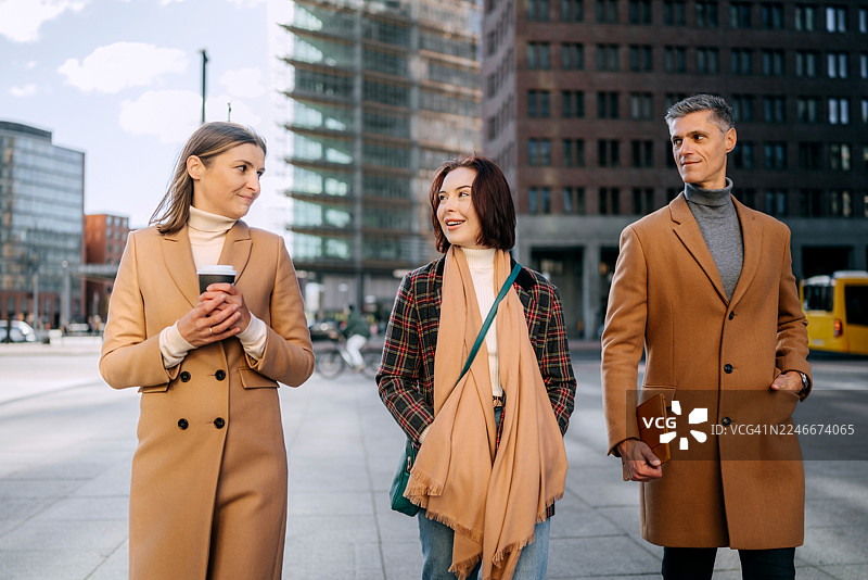 三位衣着光鲜的人在城市中行走，其中一位女士拿着咖啡杯。图片素材