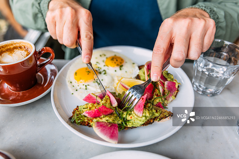 特写镜头：在咖啡馆，男人享用早餐，烤面包上铺着牛油果、萝卜和煎蛋图片素材