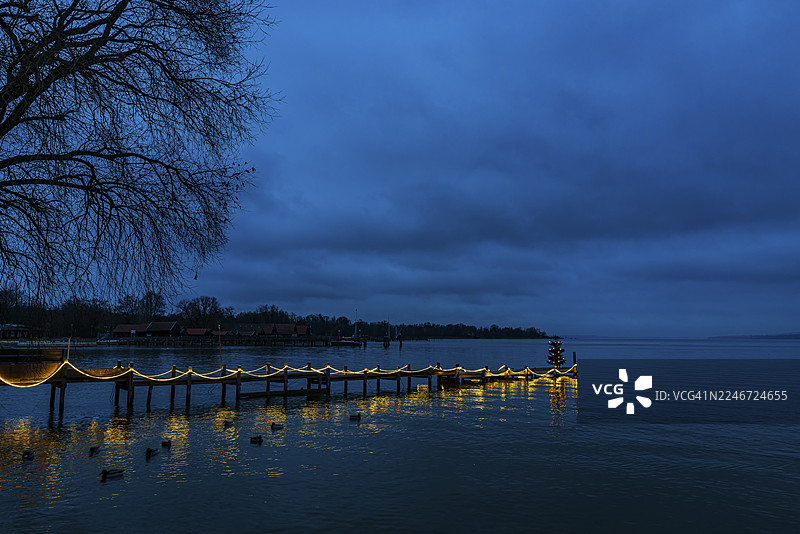 黎明时分,巴伐利亚州上巴伐利亚地区的阿默湖畔迪森,灯火通明的船坞上,一棵被照亮的圣诞树图片素材