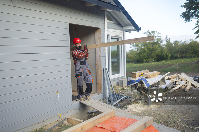 一名穿着红色安全帽的建筑工人正在一栋正在建造的木屋户外搬运木梁图片素材
