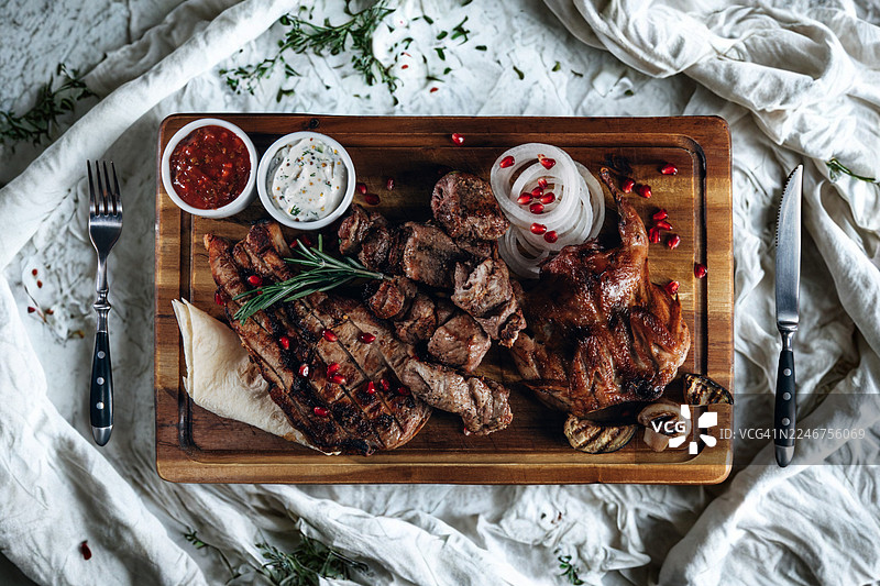 白色桌布上木板盛放的美味烤肉拼盘，俯视角度，配有酱汁、洋葱和石榴籽图片素材