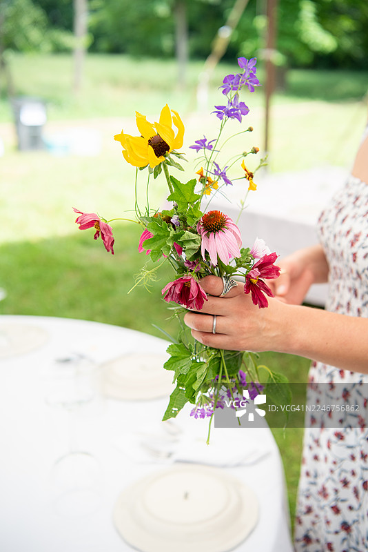 一位女士正在户外为庆祝活动准备餐桌，手中拿着一束鲜艳的野花。图片素材
