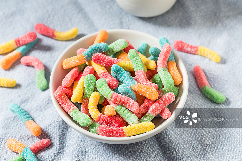 俯视彩色软糖虫糖果，放在白色碗中，碗置于浅蓝色桌布上图片素材