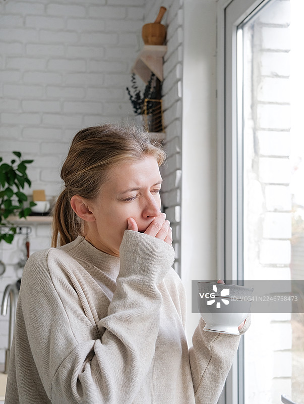 疲惫的女人在家打着哈欠，手里拿着咖啡杯，望着窗外图片素材