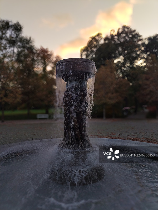 德国巴特瑙海姆公园，夕阳下的喷泉近景，水流倾泻，背景是模糊的树木。图片素材