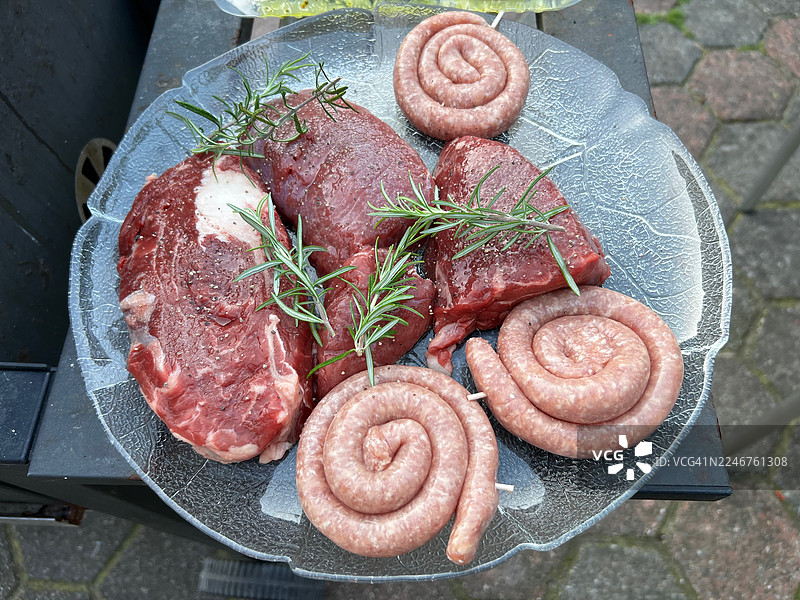 俯视角度拍摄的生肉块和香肠，用迷迭香调味，摆放在玻璃盘上，准备烧烤图片素材