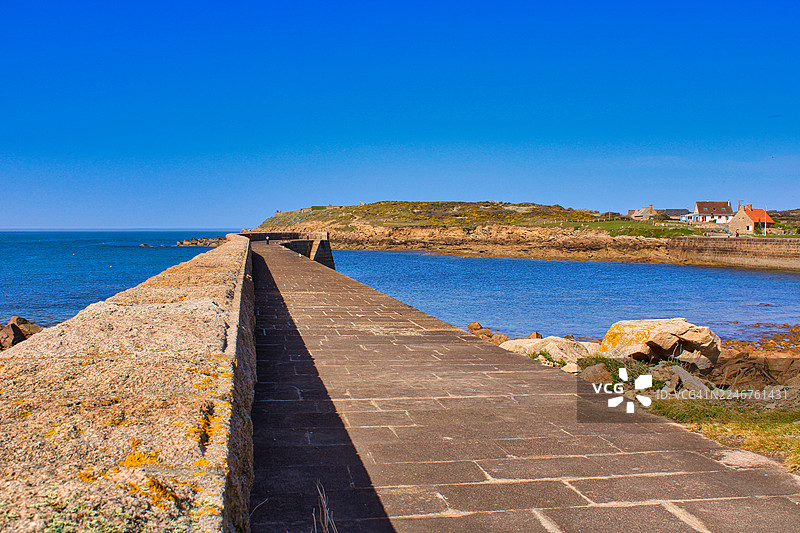 宁静的蓝色大海上一座石砌码头向海中延伸，天空晴朗，海岸线布满岩石，远处有房屋，法国费尔曼维尔图片素材