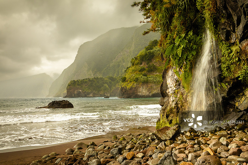 葡萄牙塞沙尔的戏剧性海岸风光，瀑布倾泻在多云天空下的岩石海滩上，海滩图片素材