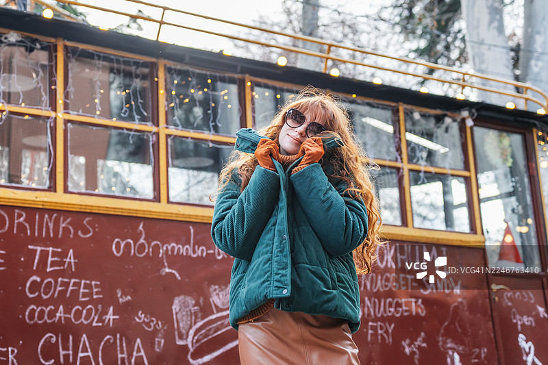 一位长着红色长发的年轻女子戴着太阳镜，穿着保暖夹克和手套，微笑着站在一个节日主题的餐车前。图片素材