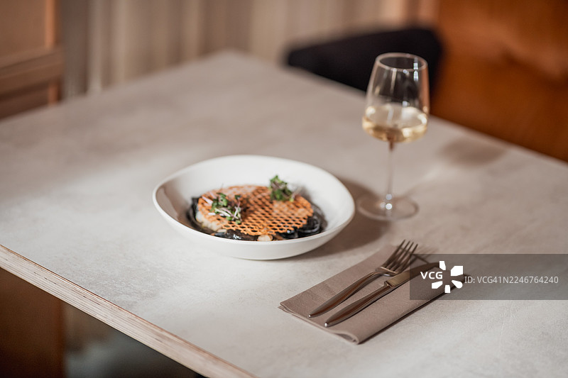 俯视角度拍摄的精致美食，配有华夫饼状的顶料和嫩芽，旁边摆放着一杯白葡萄酒、餐具，置于现代餐厅的餐桌上图片素材