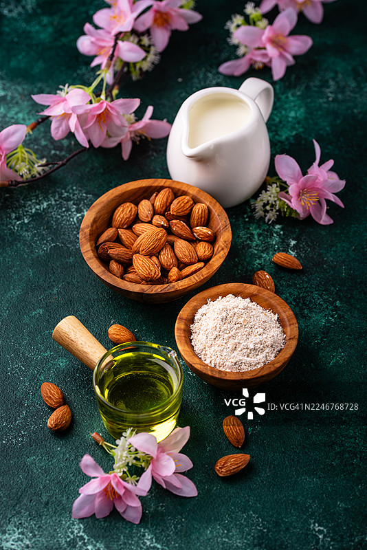 深绿色背景上的杏仁制品，包括杏仁粉、杏仁奶和杏仁油，旁边还有整颗杏仁和粉色花朵图片素材