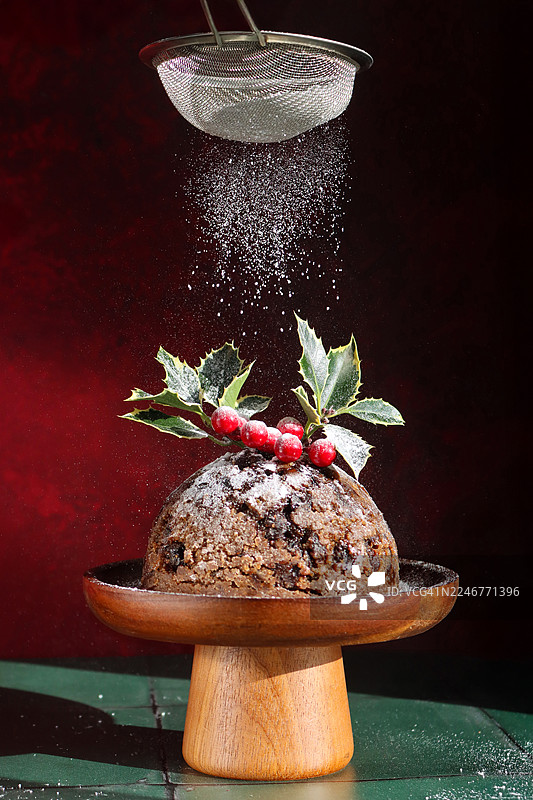 特写镜头，展示一个木制蛋糕架上放着自制的圣诞布丁，上面装饰着冬青叶和红浆果，正被一个不锈钢筛子撒上糖粉，背景为绿色表面和黑色，焦点在前景。图片素材