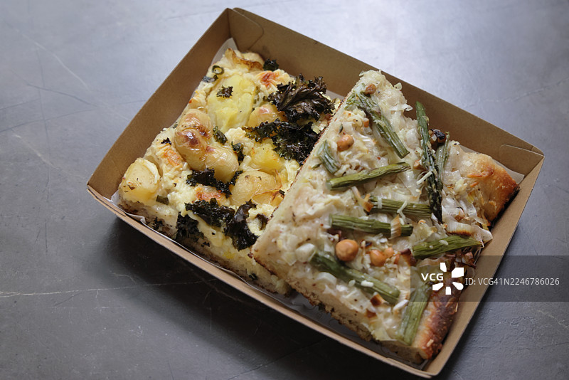 两个美味的佛卡夏面包片，一个上面有土豆、羽衣甘蓝和帕玛森芝士，另一个有芦笋、韭葱和榛子，放在纸盒外卖托盘里图片素材
