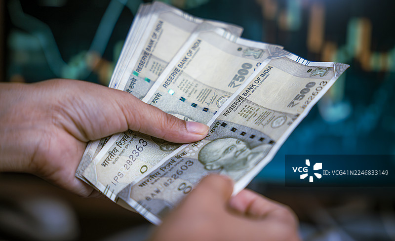 一只手拿着一叠印度卢比纸币，背景是模糊的股市屏幕图片素材