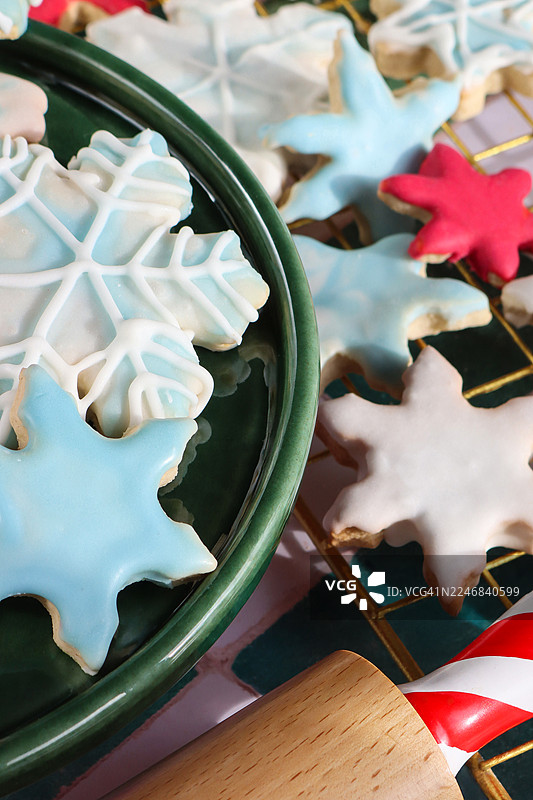 全画幅图片，展示了绿色盘子和金属冷却架上的自制圣诞点心，上面覆盖着皇室糖霜的糖饼干，有白色、红色和浅蓝色的雪花图案，还有擀面杖。背景是绿白相间的格子图案，俯视角度。图片素材