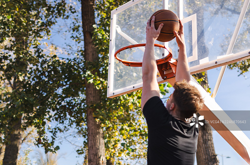 阳光明媚的白天，在树下的户外公园球场，一名篮球运动员完成了一记惊险的扣篮图片素材