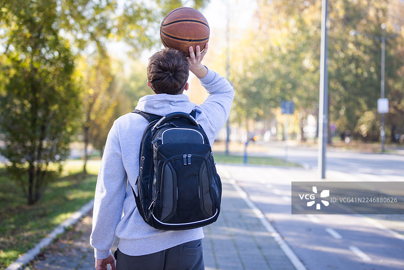 秋日阳光明媚，一名背着背包的学生在公园小径上投掷篮球图片素材
