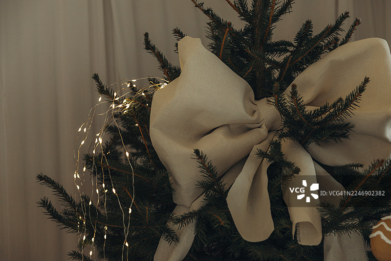 一棵圣诞树，挂着一个大大的米色蝴蝶结和串灯，靠着浅色窗帘。图片素材