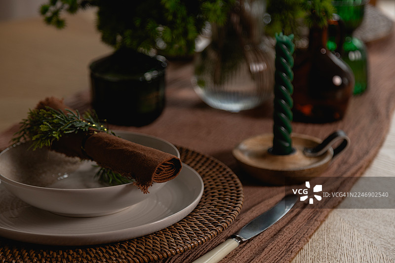 餐桌上铺着编织餐垫，上面放着一个白色碗和盘子，一条绿色枝叶固定的棕色餐巾，一个粗糙的木质烛台里放着一支深绿色扭纹蜡烛，以及玻璃瓶和花瓶里的植物。图片素材