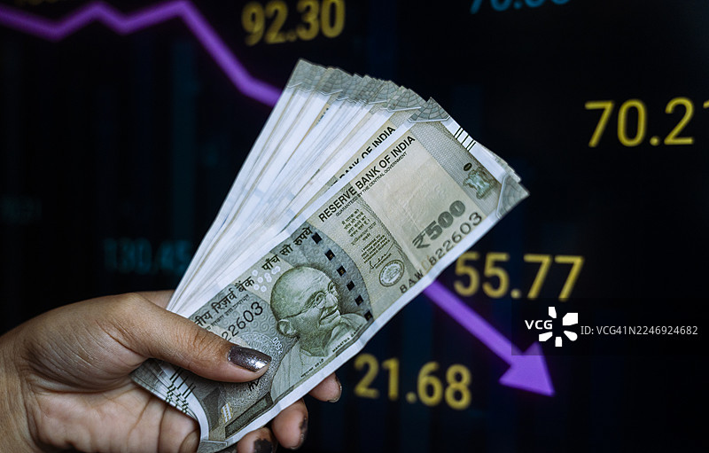 一只手拿着一叠印度500卢比纸币，背景是下跌的股市图表。图片素材