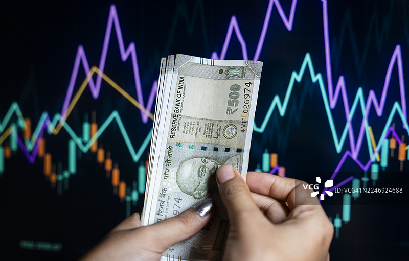 一只手拿着散开的500卢比纸币，背景是色彩斑斓的股市图图片素材