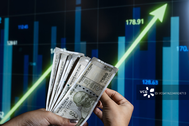 一只手拿着一叠印度500卢比纸币，背景是上升的股市图表。图片素材