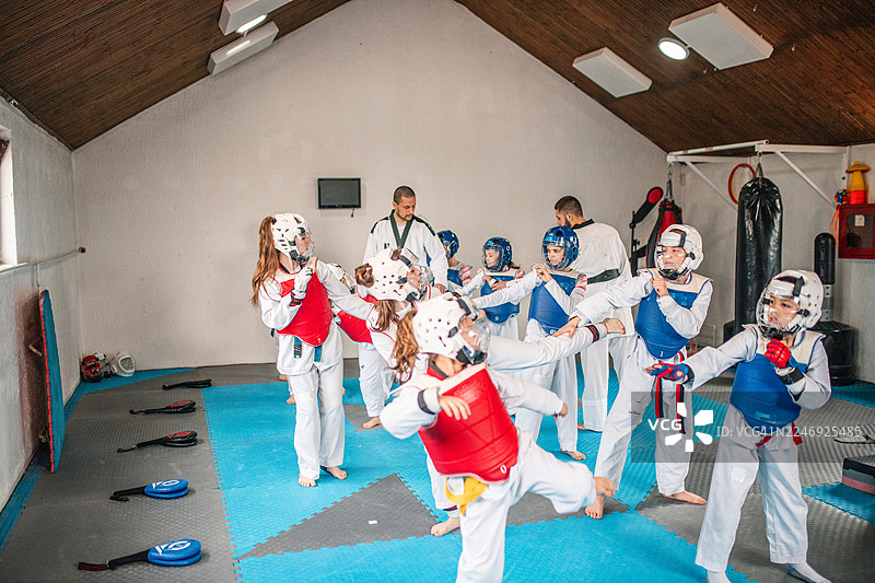 男孩和女孩们穿着跆拳道传统服饰，有的系着红腰带，有的系着蓝腰带，正在武术课上训练。两位技艺精湛的教练在旁指导他们的练习。图片素材