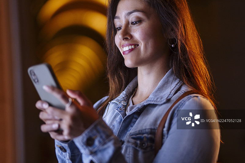 穿着休闲牛仔夹克的女士在温暖的走廊灯光下，微笑着使用智能手机图片素材