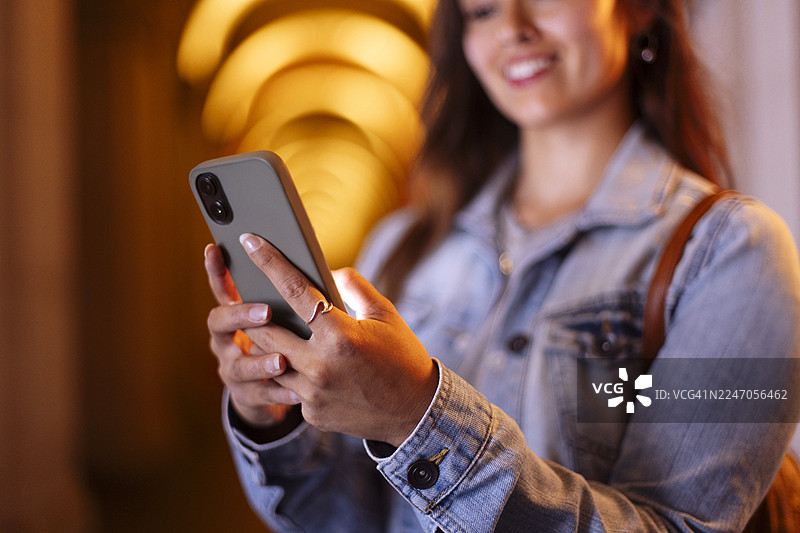 身穿牛仔夹克的女子背着背包，在温暖的走廊里微笑着使用智能手机图片素材