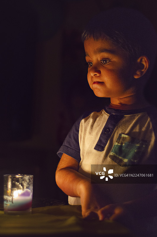 好奇/沉思 - 一个深色头发的小男孩正专注地低头看着玻璃烛台里摇曳的蜡烛，在黑暗的环境中，他的脸被温暖的光芒柔和地照亮。这张照片营造出一种奇妙、沉思或安静专注的时刻感。图片素材