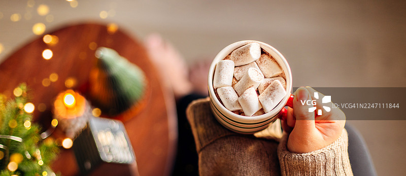 女性双手捧着温暖的热巧克力杯，杯中放着棉花糖，身穿舒适的毛衣，在家中。节日装饰，营造出舒适的冬日氛围。图片素材