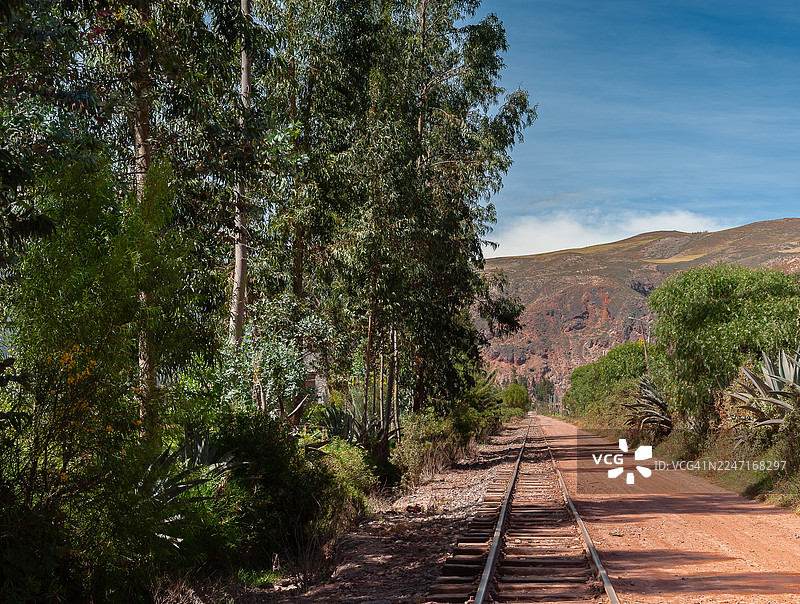 空旷的铁轨和一条蜿蜒穿过树林的土路，背景是山脉，天空湛蓝，乌鲁班巴，秘鲁图片素材