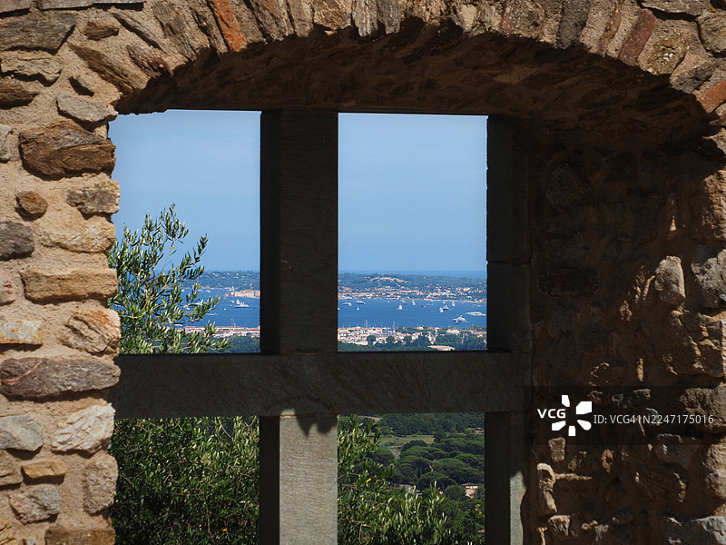 阳光明媚的一天，透过古老的石头窗框，可以看到法国格里莫海滨小镇和海上的船只的风景。图片素材