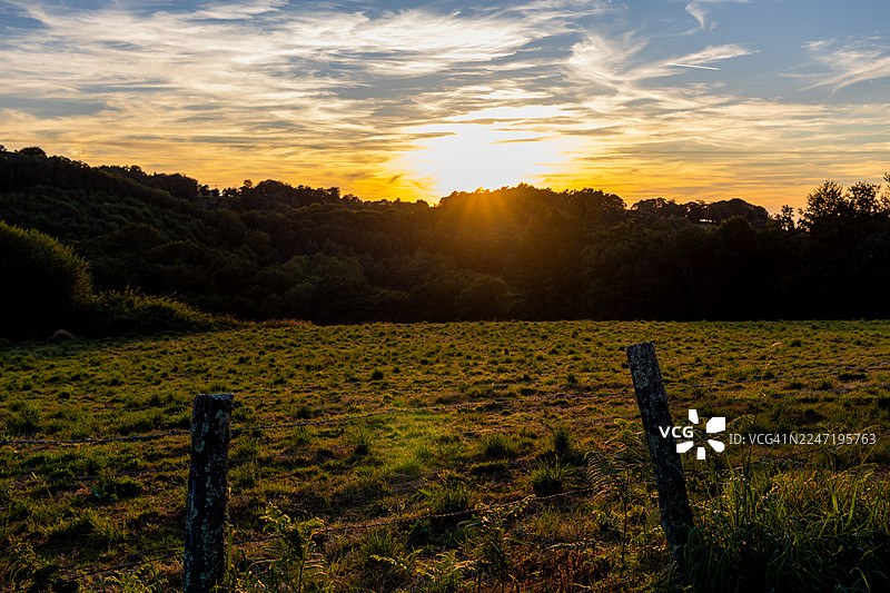 夕阳西下时，郁郁葱葱的绿野与绚丽的天空构成了一幅美景，阳光透过树林洒下光芒。图片素材