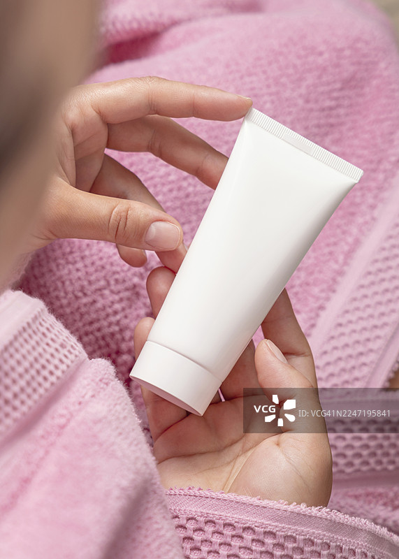 女子在家中涂抹保湿霜的特写，粉色毛巾背景，手中拿着一个空白的白色管状瓶图片素材