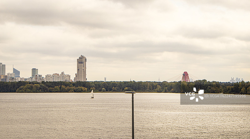 广阔河流上横跨一座红色大桥的现代城市景观全景，天空多云图片素材