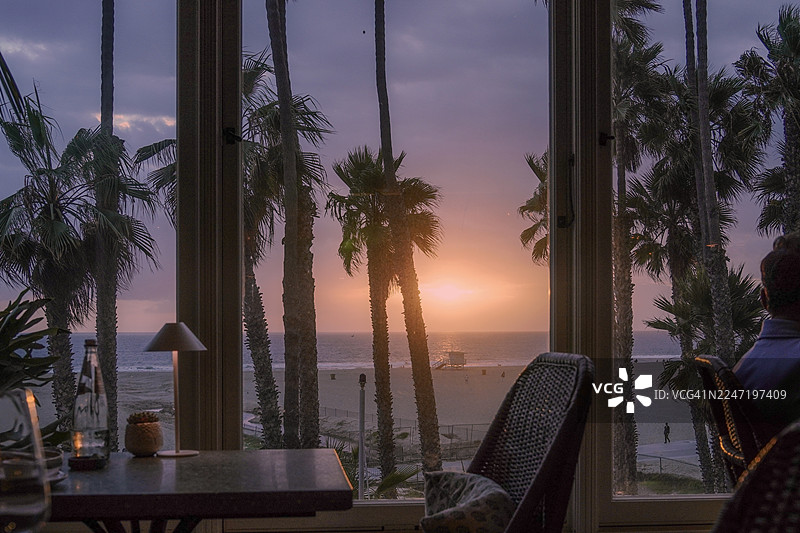 日落时分，透过窗户看到的壮丽海景与天空，窗外有棕榈树，还能看到一个人的一部分身影图片素材