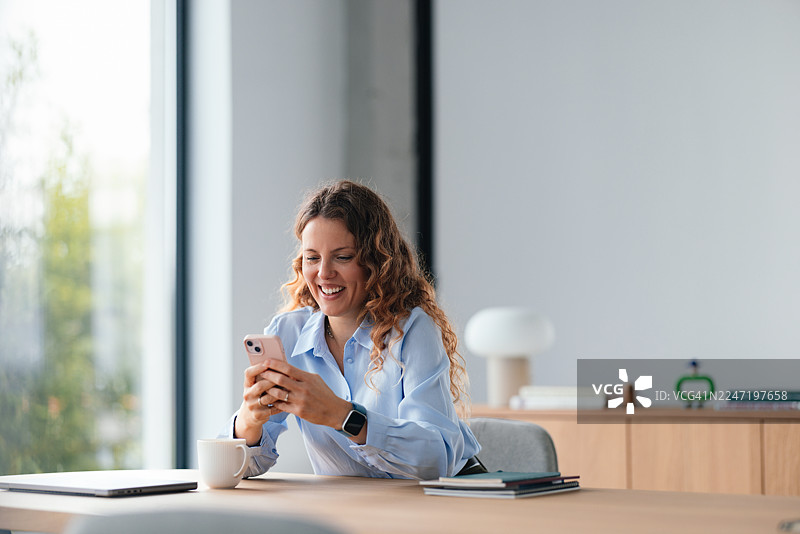 一位面带微笑的职业女性在明亮的办公空间里，于高效工作日的早晨，在现代办公桌前使用智能手机。图片素材