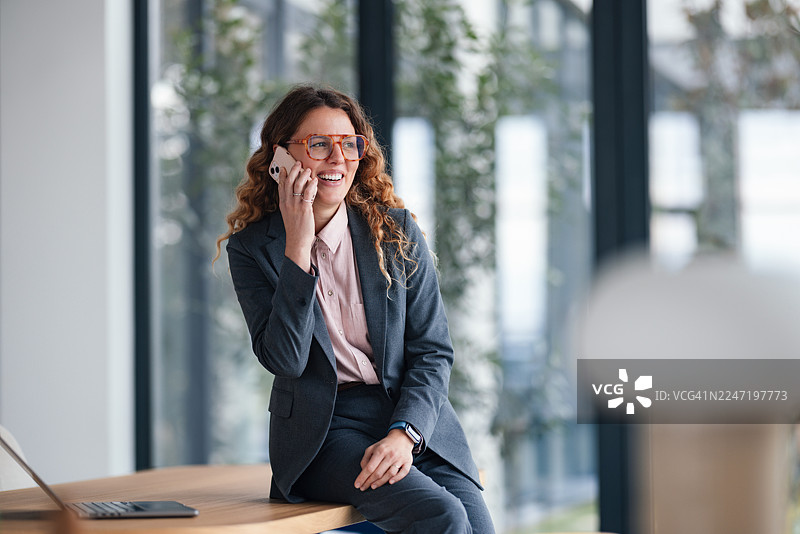 职业女性在现代办公室的办公桌旁，手持电话，旁边放着笔记本电脑图片素材