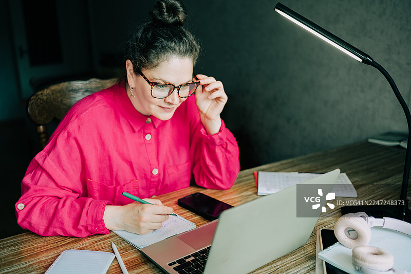 一位成熟的成年女性正在使用笔记本电脑在线学习并做笔记图片素材
