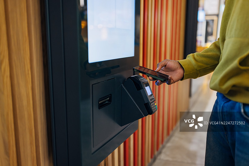 一名男子在现代快餐店使用智能手机在自助点餐机上完成食品购买，身份无法识别图片素材