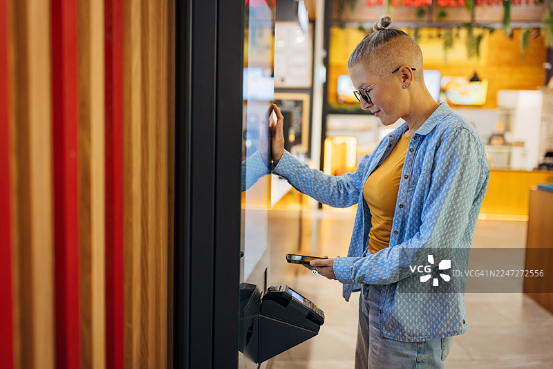 一位女性在现代快餐店使用智能手机在数字售货亭完成食品购买图片素材