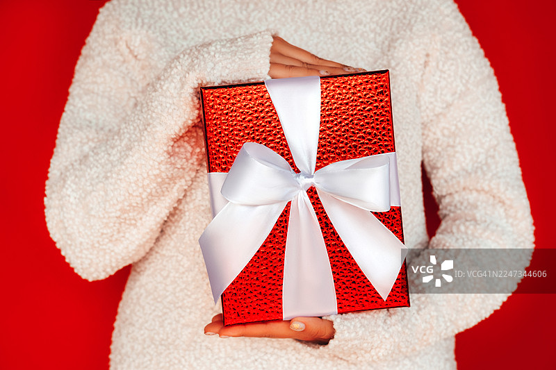 穿着柔和波点羊毛毛衣的女性，手持带有白色蝴蝶结的亮红色礼品盒，背景为红色。圣诞和新年庆祝概念。正面视角。图片素材