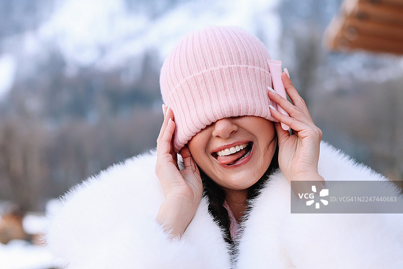冬季旅行中，女孩在雪山背景下，拨弄着粉色帽子，开怀大笑，展现出俏皮的表情，充满趣味的肖像，概念为玩耍图片素材